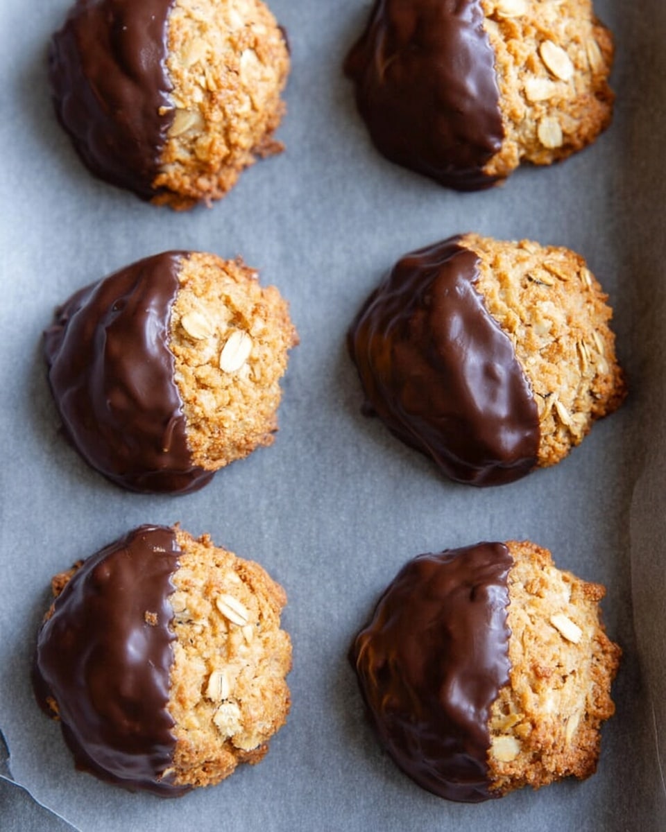 The image shows six round cookies on a gray baking tray lined with parchment paper. Each cookie has one half covered in shiny dark chocolate while the other half is light golden brown with a rough and crunchy texture, showing oats and a slightly uneven surface. The cookies are arranged in two rows of three, all spaced evenly apart. Photo taken with an iphone --ar 4:5 --v 7