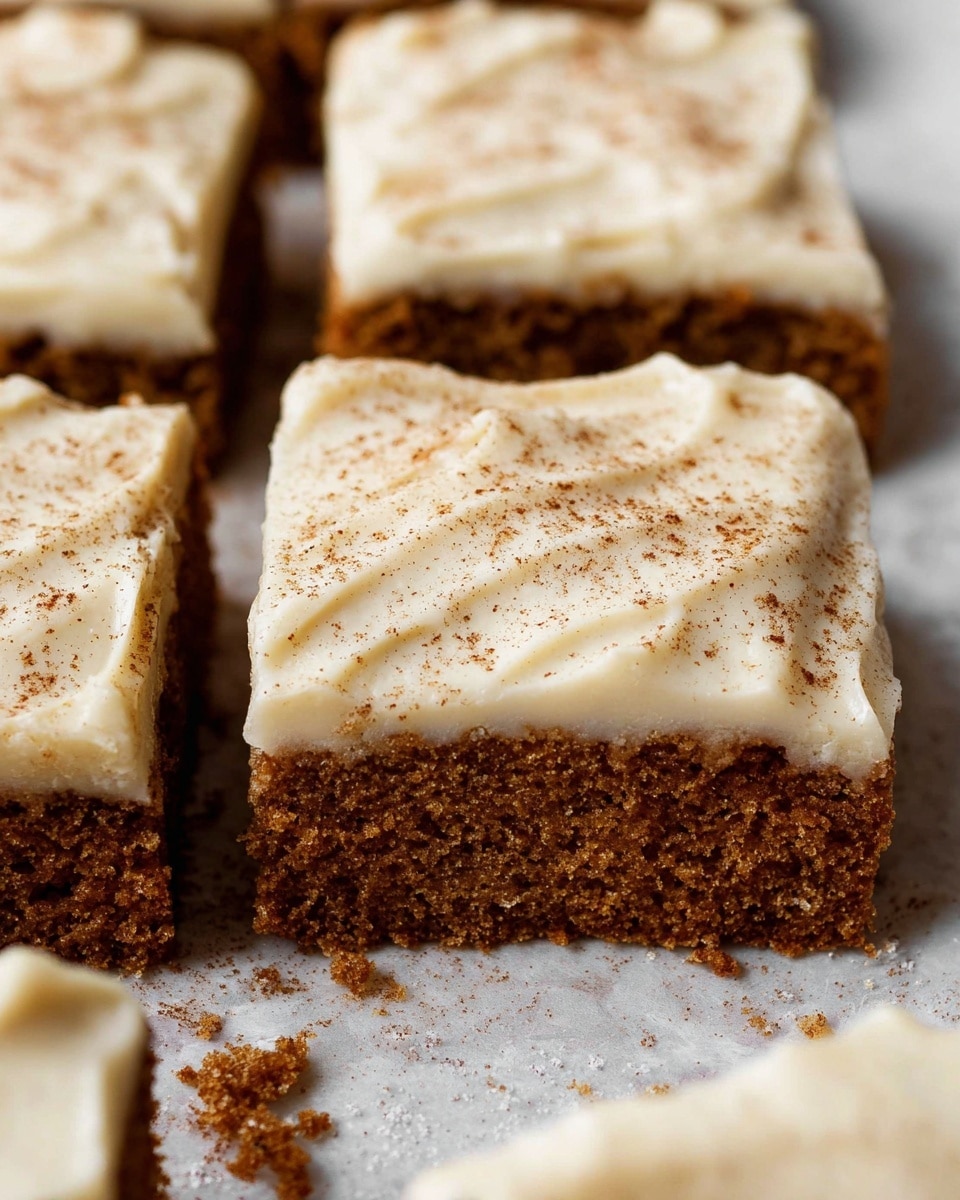 A close-up view of cut square pieces of moist brown cake with a thick, creamy off-white frosting layer on top, slightly textured and sprinkled lightly with cinnamon powder. The cake layer looks soft with small air holes, and the frosting is spread smoothly but shows gentle swirls and folds. The cake squares rest on a white marbled textured surface with a few crumbs scattered around. photo taken with an iphone --ar 4:5 --v 7