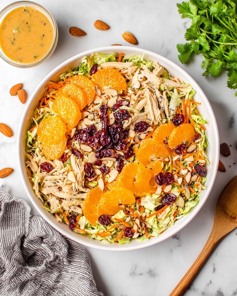 A large white bowl filled with a colorful salad sitting on a white marbled surface, with shredded light green cabbage and orange grated carrots forming the base layer. On top, there are bright orange mandarin slices scattered evenly along with pieces of shredded chicken, dark red dried cranberries, and thinly sliced almonds adding texture. Small black poppy seeds are sprinkled over the top, giving a speckled look. To the left, there is a small glass of light orange dressing, and a bunch of fresh green cilantro lies in the upper right corner. A wooden spoon is placed partly under the bowl, and a gray and white striped cloth is partially visible at the bottom left corner. Photo taken with an iphone --ar 4:5 --v 7