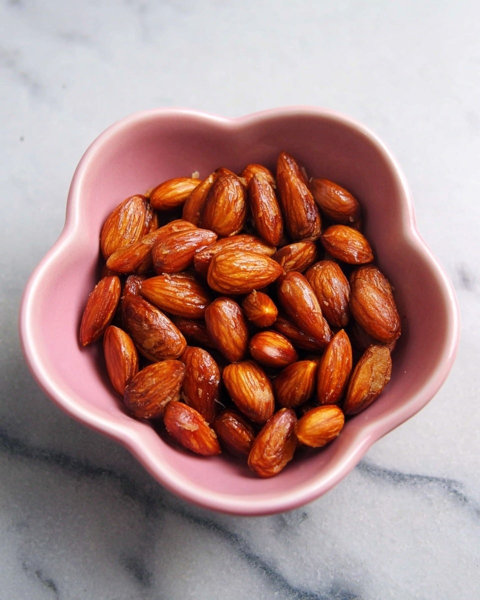 A pink bowl shaped like a flower holds a single layer of roasted almonds. The almonds are shiny with a rich brown color, showing slight variations from light to dark brown, and a smooth roasted texture. The bowl sits on a white marbled surface, creating a clean and bright background that contrasts with the warm tones of the almonds. photo taken with an iphone --ar 4:5 --v 7