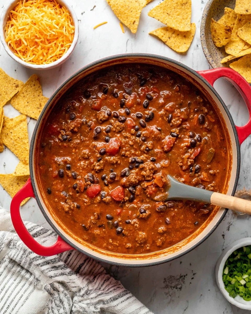 A white bowl filled with thick chili showing layers of brown beans and ground meat in a rich red sauce. On top of the chili is a layer of melted yellow cheese sprinkled with small pieces of chopped green onions. Three triangular yellow corn chips are placed inside the bowl, resting on the chili near the edge. A black spoon stands inside the bowl on the right side, touching the chili. Around the bowl, there are more yellow corn chips scattered on a white marbled surface. A white cloth with black stripes is partially under the bowl, and small wooden bowls with shredded yellow cheese and chopped green onions sit in the background. Photo taken with an iphone --ar 4:5 --v 7
