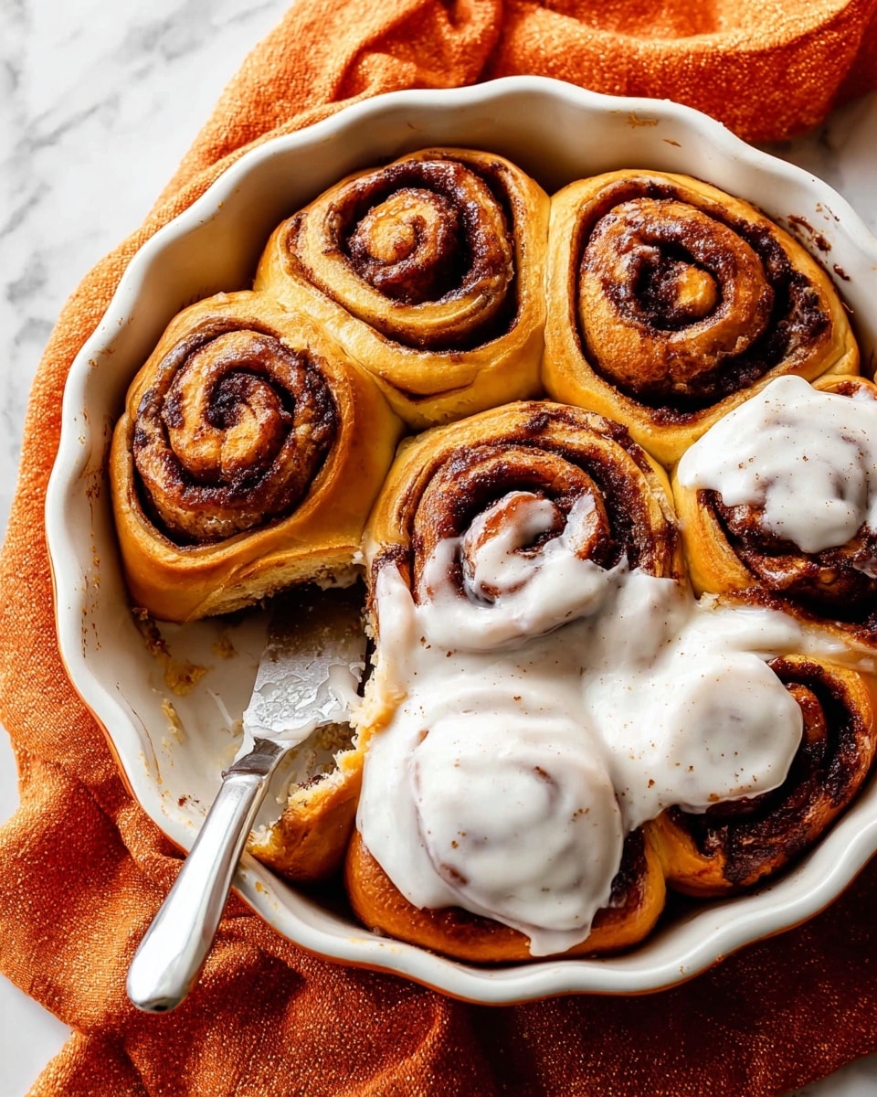 This image shows a round batch of seven cinnamon rolls baked in a white ceramic dish with a scalloped edge. The rolls have a golden brown dough base with swirls of dark cinnamon filling visible in each one, creating a spiral pattern. Four of the rolls have thick, smooth white icing with tiny specks, spread over their tops, while the other rolls are bare, showing their shiny, slightly caramelized texture. A silver spreading knife with white icing on it rests on one of the iced rolls. The dish sits on an orange cloth on a white marbled surface. Photo taken with an iphone --ar 4:5 --v 7