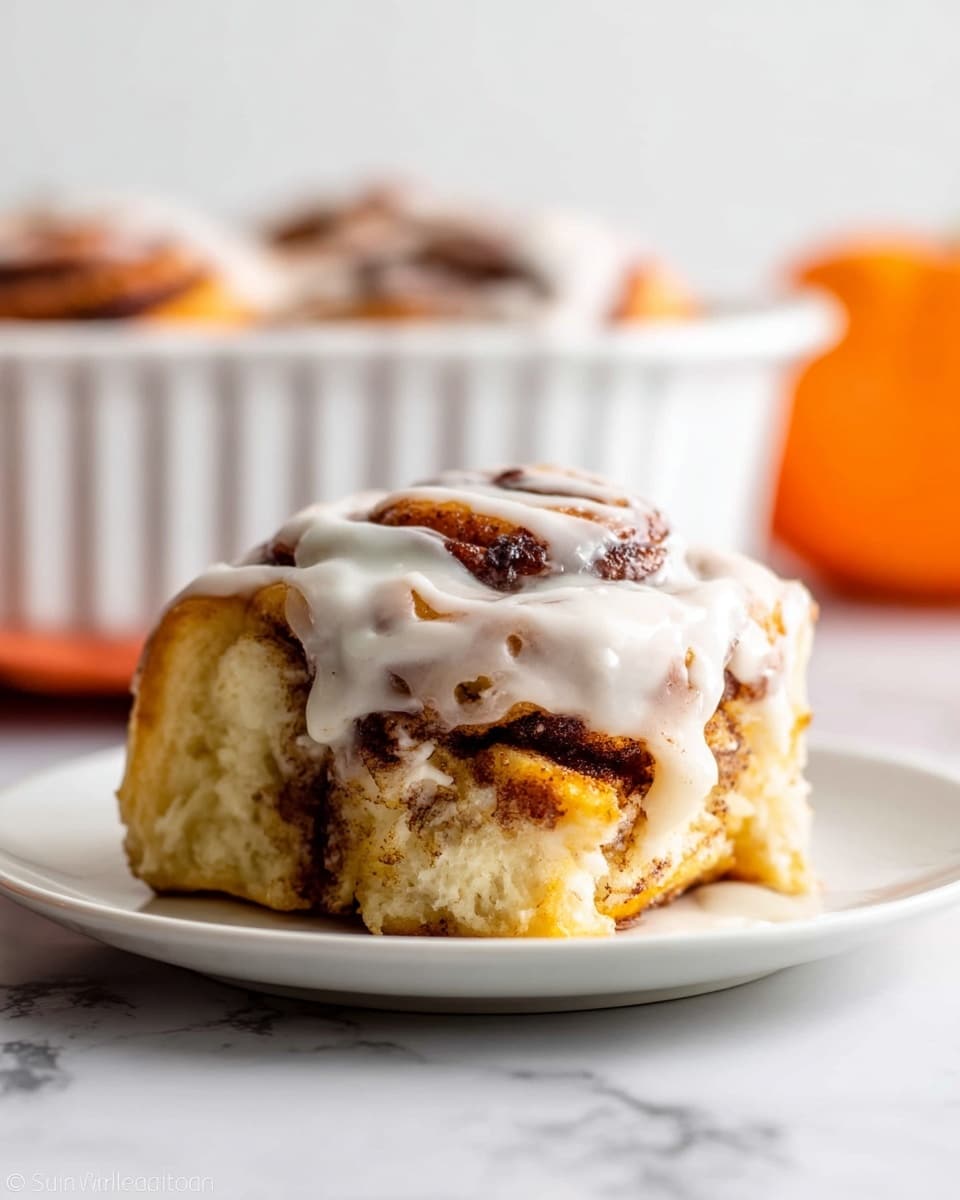A close-up of a single cinnamon roll on a white plate with three visible layers: the bottom layer is soft and golden brown dough, the middle layer is swirled with dark cinnamon filling, and the top layer is thick, creamy white icing that drips down the sides. In the background, a blurred white fluted dish holds more cinnamon rolls, all set on a white marbled surface with an out-of-focus orange object on the side. photo taken with an iphone --ar 4:5 --v 7