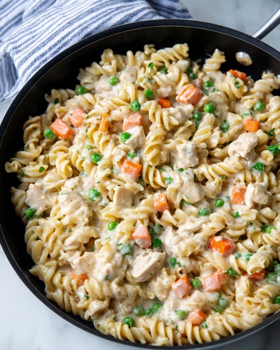 A close-up view of a black pan filled with creamy rotini pasta mixed with small pieces of tender white chicken, bright orange carrot cubes, and fresh green peas, all coated evenly in a smooth, white sauce. The pan is placed on a white marbled surface with a hint of a striped cloth underneath. photo taken with an iphone --ar 4:5 --v 7