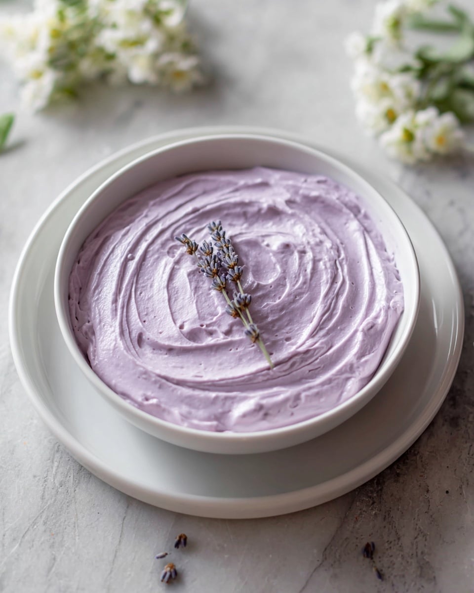 A single-layer dessert in a white bowl shows a smooth, thick, light purple creamy layer spread evenly with soft swirls across the top. There is a small sprig of dried lavender placed in the center as a simple decoration. The bowl sits on a matching white plate on a white marbled surface. Some blurred white and green flowers surround the bowl, adding a gentle natural touch to the scene. Photo taken with an iphone --ar 4:5 --v 7