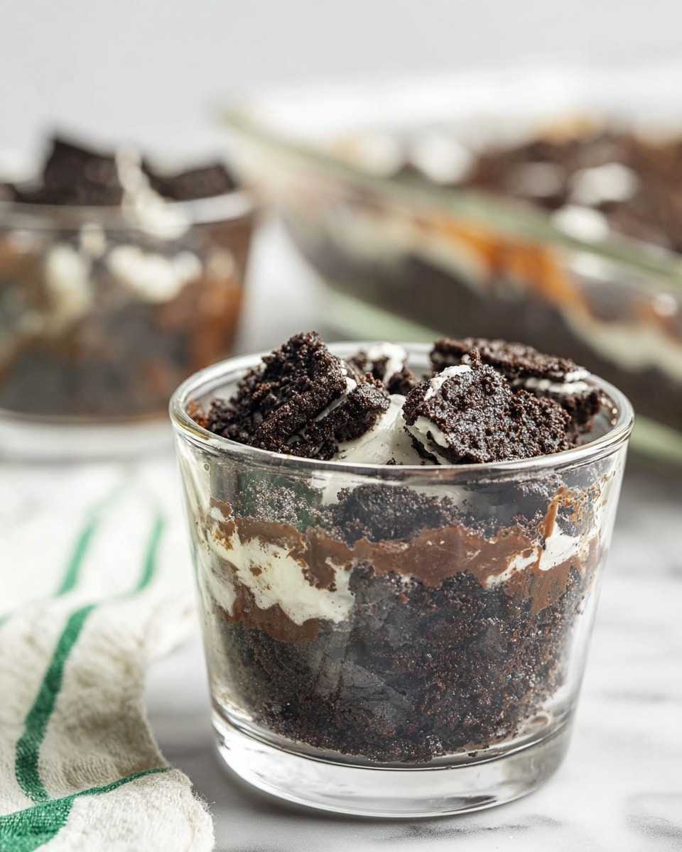 The image shows a small clear glass filled with a dessert that has two layers. The bottom layer is dark brown and crumbly, looking like crushed chocolate cookies mixed with some moist filling. The top layer is pieces of chocolate cookies with white cream visible on some pieces, placed unevenly. In the background, there is a blurred view of another similar glass filled with the same dessert and a glass baking dish with more of the dessert inside. The setting has a white marbled texture surface with part of a white and green striped cloth on the left side. Photo taken with an iphone --ar 4:5 --v 7