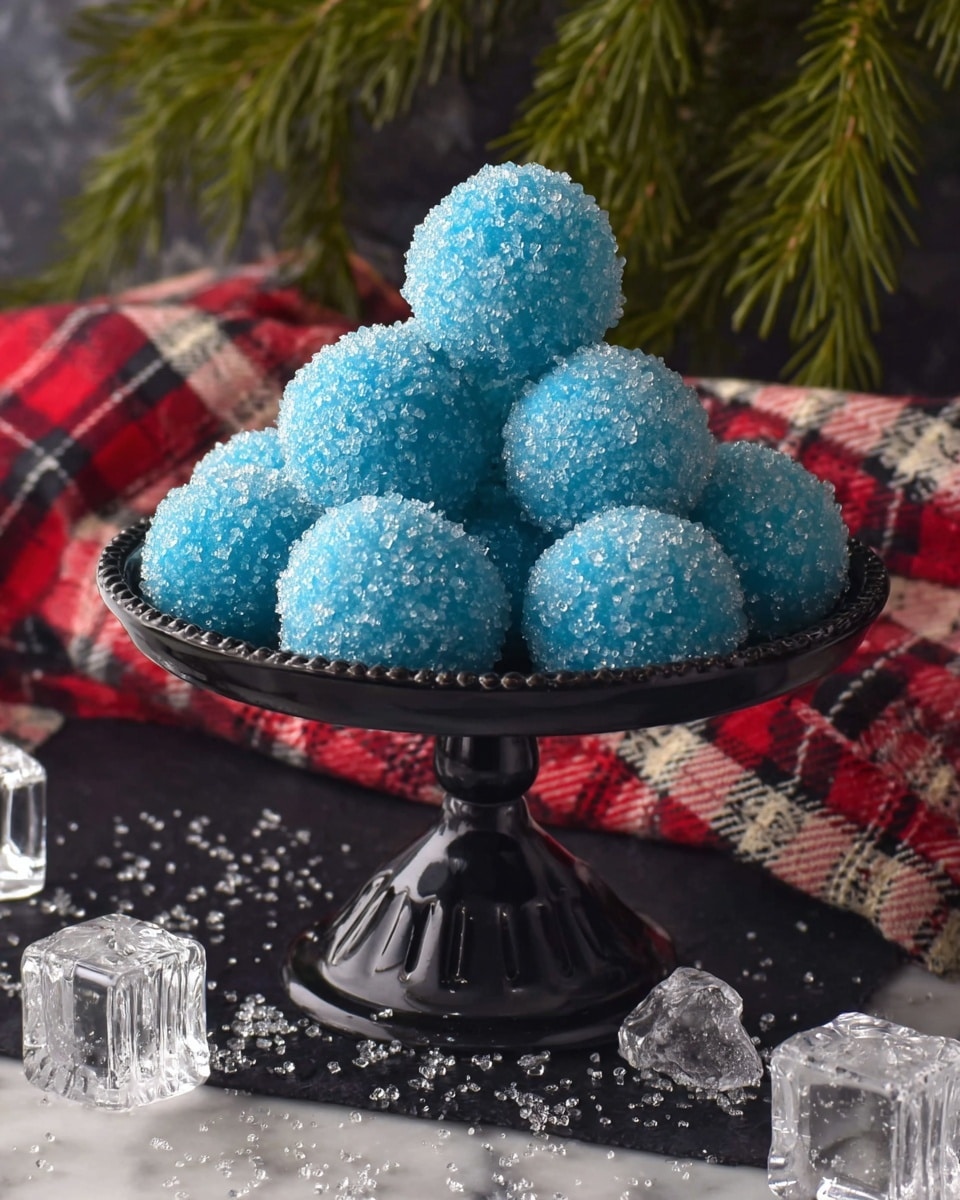 A black metal pedestal holds a pyramid stack of bright blue spherical candies coated fully with large sparkling sugar crystals. The candy balls appear smooth under the crystals and are tightly packed in three visible layers, with the top layer showing a single ball, the middle layer with four, and the bottom layer with five balls. The stand sits on a dark cloth sprinkled with scattered large sugar crystals and clear plastic gem decorations shaped like ice cubes and beads. Behind the pedestal, there is a white marbled textured surface with a red and cream plaid cloth slightly visible, along with green pine branches adding a festive touch. Photo taken with an iphone --ar 4:5 --v 7