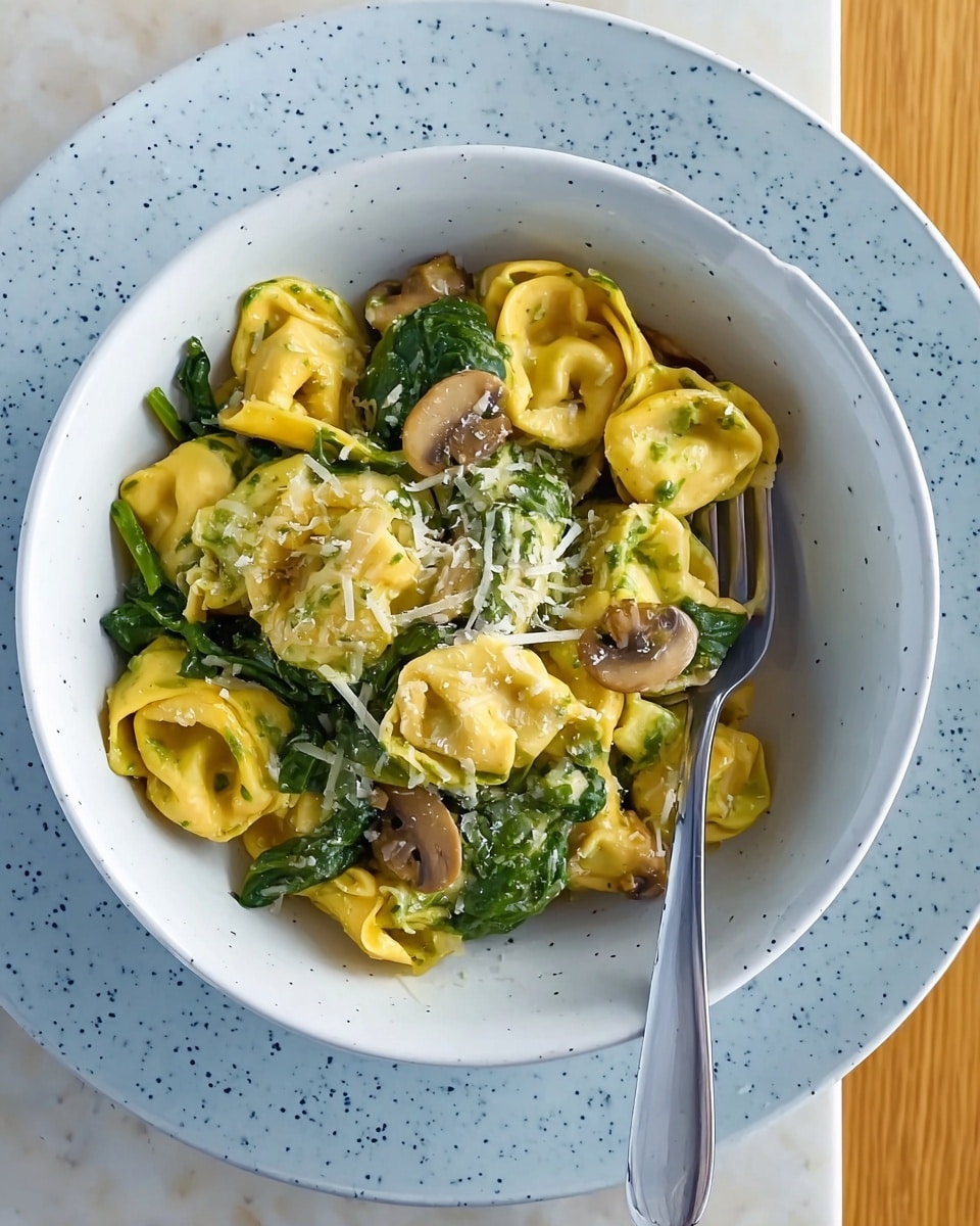 A bowl filled with two-layered tortellini pasta, the outer layer bright yellow and the inner layer light green, showing their folded and stuffed shapes. The tortellinis are mixed with vibrant green spinach leaves and thin slices of sautéed mushrooms with a glossy, slightly brown hue. Shredded white cheese is sprinkled on top, adding texture and a soft contrast to the pasta. The bowl is white with light blue speckles and rests inside a larger matching white speckled plate. A silver fork sits on the plate's right side, and the whole setting is placed on a white marbled surface. photo taken with an iphone --ar 4:5 --v 7