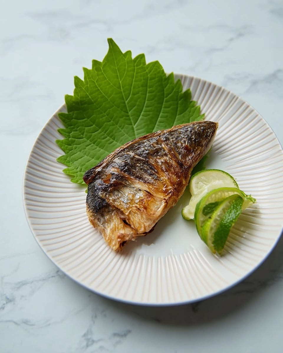 A single piece of grilled fish with a shiny, caramelized brown top layer and light beige, tender bottom layer sits in the center of a white plate with speckled texture and a thick rim. Behind the fish, there is a dark green shiso leaf with jagged edges, and a small wedge of light green lime is placed next to the fish on the right side. The plate rests on a white marbled surface with a cream-colored cloth visible in the background. Photo taken with an iphone --ar 4:5 --v 7