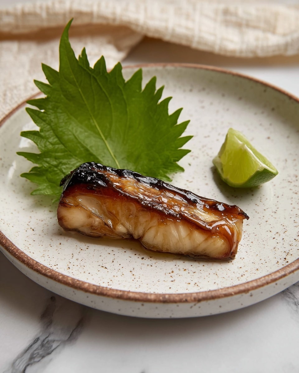A single piece of golden-brown grilled fish with crispy darkened skin lies in the center of a white plate with a ridged design. Behind the fish, there is a large, textured green leaf that adds a fresh look. To the right of the fish, a small, twisted slice of pale green citrus peel curls upwards next to a chunk of translucent light green lime. The plate sits on a white marbled surface. photo taken with an iphone --ar 4:5 --v 7