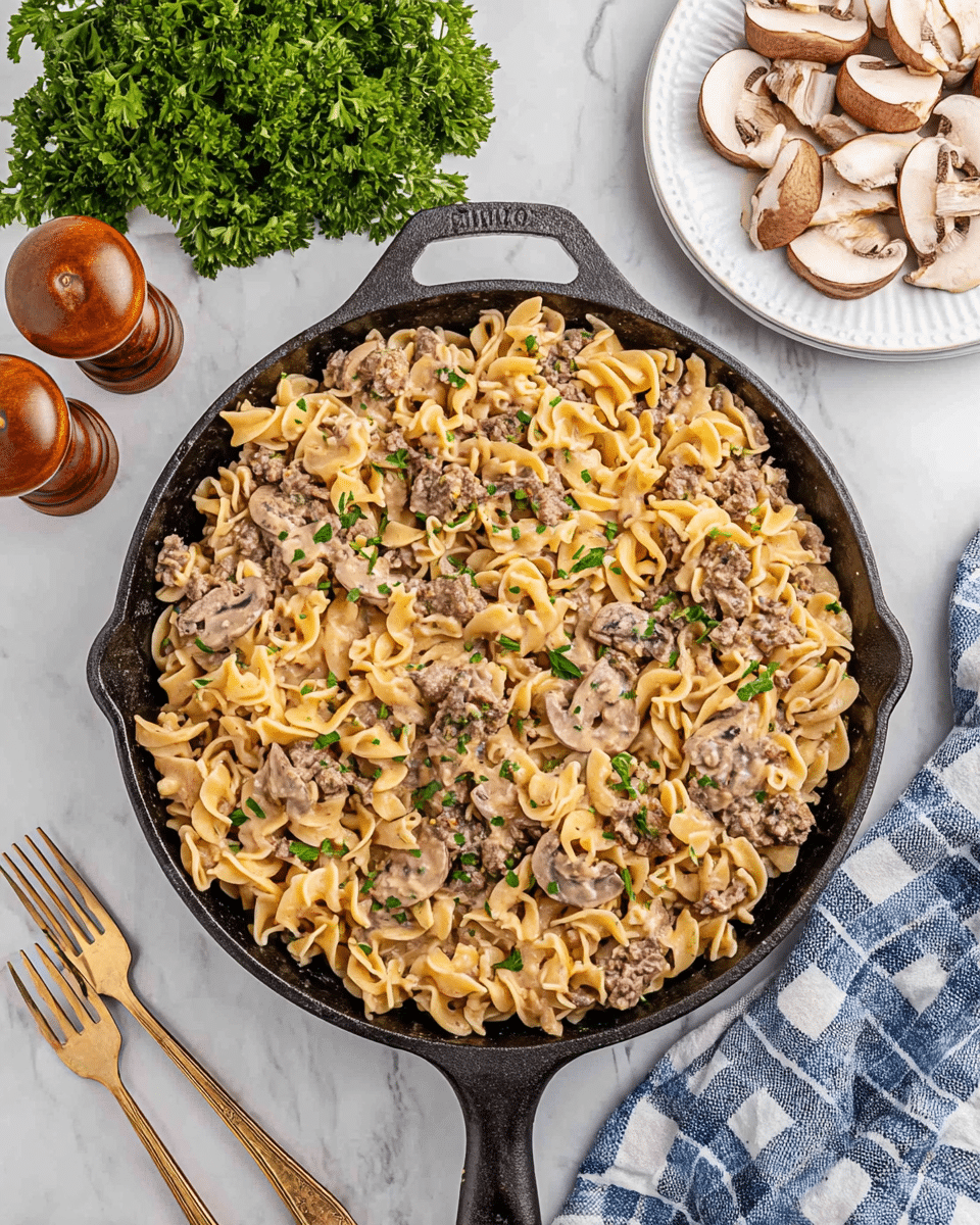 A black cast iron skillet filled with creamy pasta made of short twisted noodles mixed with ground meat and sliced brown mushrooms, all coated in a light beige sauce, topped with small green parsley leaves for garnish. The skillet sits on a white marbled surface. Around the skillet, there is a bunch of fresh green parsley on the top left, two wooden brown salt and pepper shakers above, three sliced mushrooms on the top right, a white plate with two forks on the bottom left, and a white cloth with blue checkered pattern on the bottom right. Photo taken with an iphone --ar 4:5 --v 7