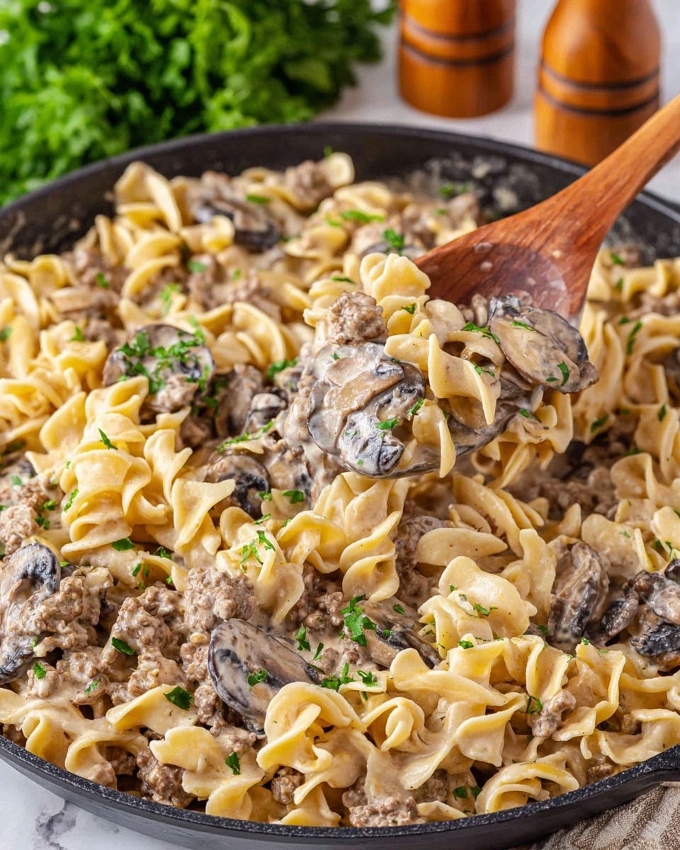 A close-up view of a creamy pasta dish served in a black skillet. The dish has three main layers: at the base, there are many egg noodles that are pale yellow and soft, twisted into irregular shapes. Mixed evenly throughout the noodles are slices of dark brown mushrooms with a smooth texture and chunks of light brown ground meat. The entire dish is coated in a light beige creamy sauce that looks smooth and slightly thick. Small bits of fresh green parsley are sprinkled on top as a garnish. A wooden spoon is lifting a portion of the pasta near the center left, showing the noodles, mushrooms, and meat clearly. In the background, some green leafy herbs and wooden salt and pepper shakers are blurred. The surface is a white marbled texture. photo taken with an iphone --ar 4:5 --v 7