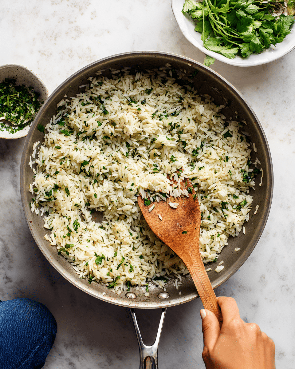 A gray pan filled with cooked rice mixed evenly with small green herb pieces, giving the rice a light green color and textured look. A woman's hand is holding a wooden spatula, stirring the rice near the bottom right corner. The pan is placed on a white marbled surface, with a white bowl on the top right containing fresh green leafy herbs and other ingredients visible. Part of a person's blue jeans can be seen at the bottom edge of the image. photo taken with an iphone --ar 4:5 --v 7