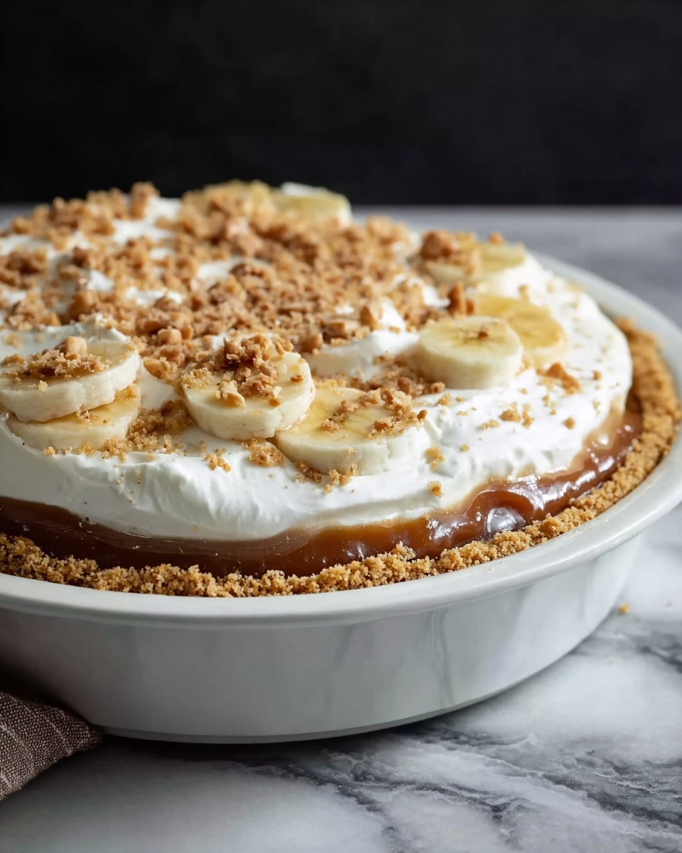 A white pie dish holding a three-layer dessert starting with a golden brown crumb crust that forms the base and edges. Above the crust is a thick layer of smooth, shiny caramel or toffee sauce topped with round slices of pale yellow bananas partly visible under a thick, fluffy white whipped cream layer. The top is sprinkled with small pieces of crushed nuts or toffee bits scattered unevenly. The dish sits on a white marbled surface with a dark background. photo taken with an iphone --ar 4:5 --v 7