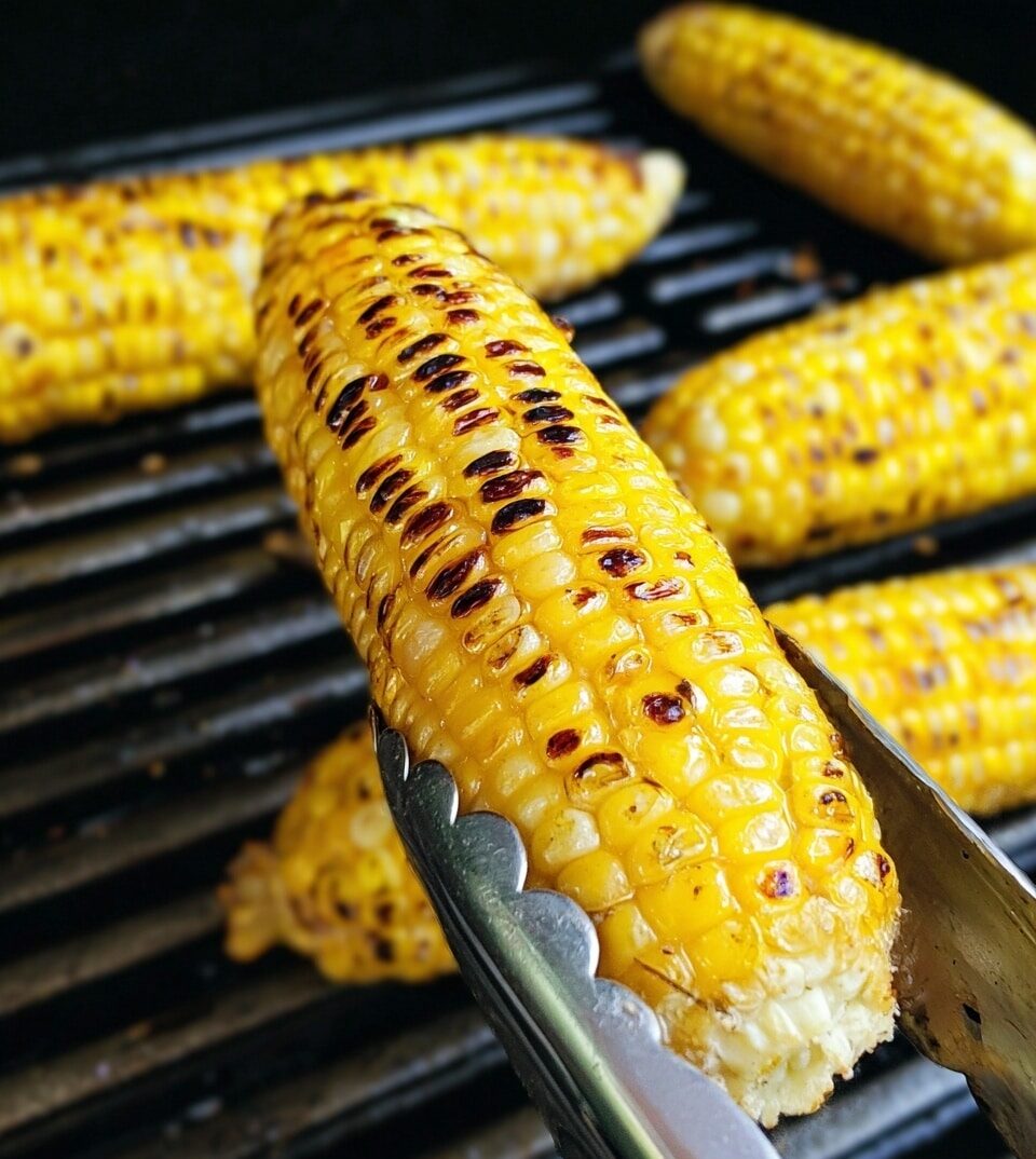 A close-up view of a yellow corn ear being held with metal tongs, showing dark brown grill marks scattered across the kernels on the top and middle layers, while the corn beneath has a mixture of bright yellow and pale yellow kernels. In the background, more ears of corn with similar grill marks rest on a dark grill surface. photo taken with an iphone --ar 4:5 --v 7