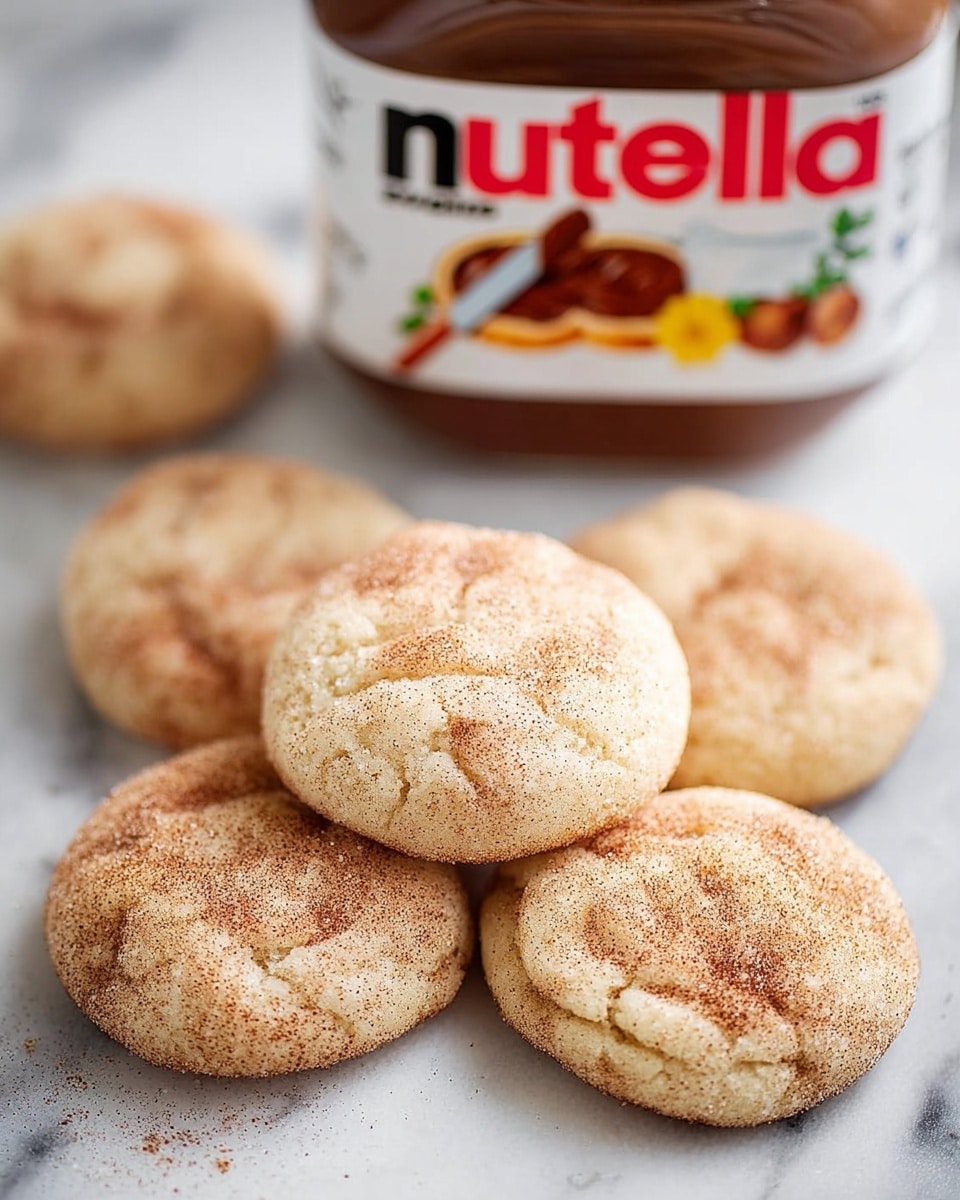 A close-up view of six round, soft cookies with a light golden-brown color and a slightly cracked surface, dusted with cinnamon and sugar. The cookies are placed directly on a white marbled texture. In the background, there is a clear jar of Nutella with a white lid, showing a label with red and black text and images of hazelnuts and a spread knife. The jar is slightly out of focus, emphasizing the cookies in the foreground. Photo taken with an iphone --ar 4:5 --v 7