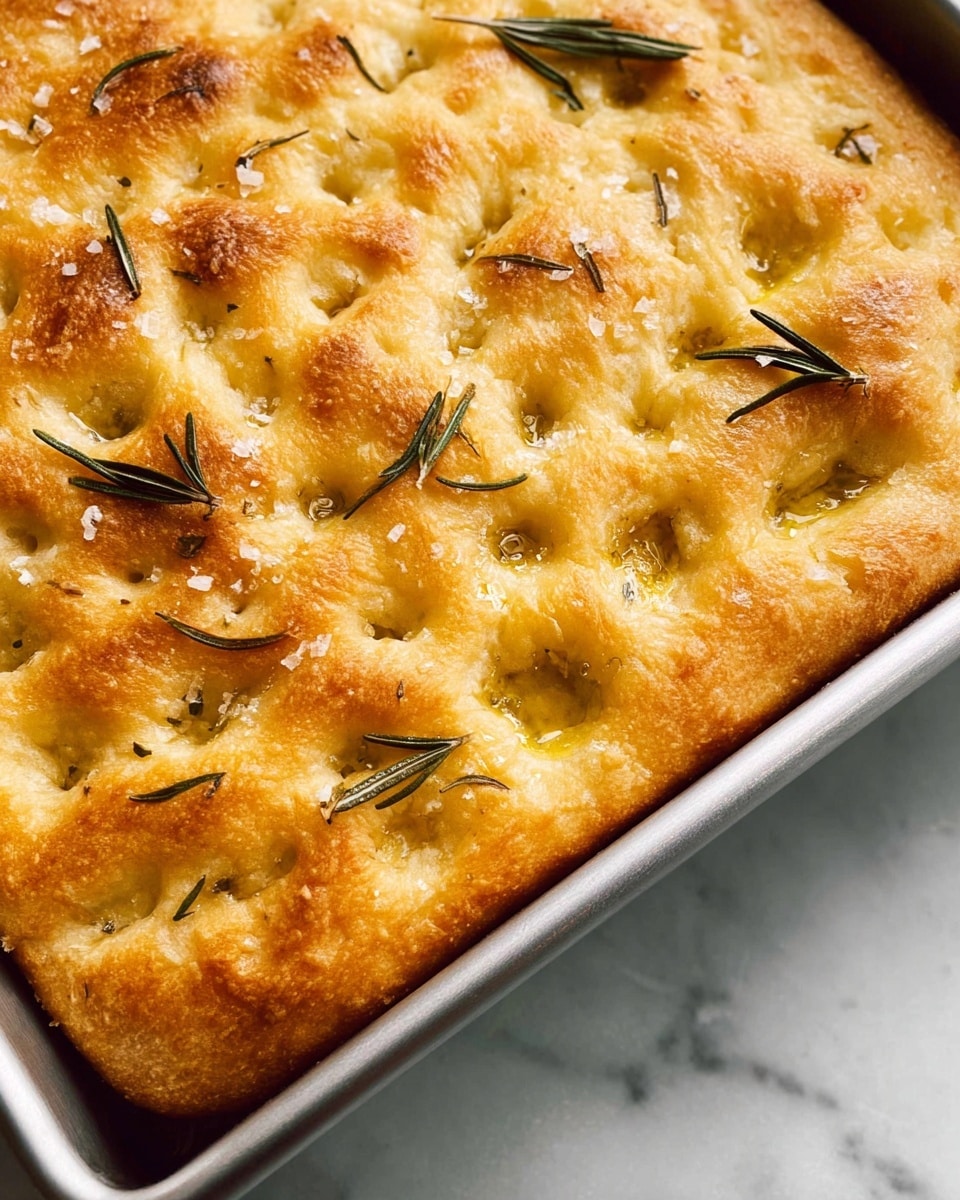 The image shows a close-up of a golden-baked focaccia bread in a metal rectangular pan, featuring a textured surface with small dimples and a light crust. The bread’s top layer is pale yellow with a glossy finish, scattered with fresh rosemary leaves and coarse sea salt crystals, adding texture and a hint of green contrast. The edges are slightly raised and crisp, framing the soft and airy interior visible near the corners. The pan sits on a white marbled surface, enhancing the warm tones of the bread. photo taken with an iphone --ar 4:5 --v 7