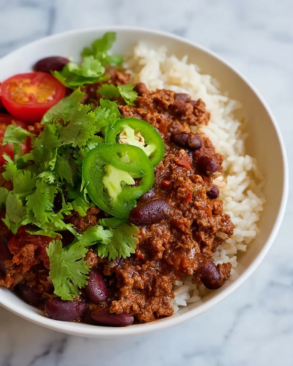A white bowl is filled with a base layer of cooked white rice, showing individual grains with a soft texture. On top of this is a thick layer of chili made from ground meat and dark red kidney beans mixed in a rich, brownish-red sauce. The chili is garnished with bright green cilantro leaves spread across, slices of fresh green jalapeño pepper placed near the center, and halved cherry tomatoes adding a burst of red color. The bowl is placed on a white marbled surface. Photo taken with an iphone --ar 4:5 --v 7