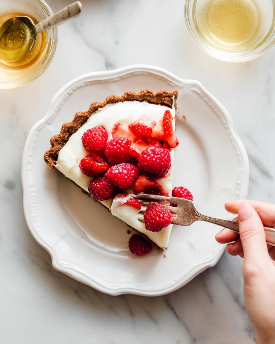 A slice of tart sits on a white scalloped plate on a white marbled surface; the tart has a browned crust at the edge, topped with a thick layer of smooth white cream, and fresh bright red raspberries and sliced strawberries scattered on top. A woman's hand holds a fork piercing the tart near the fruit, ready to take a bite. To the side, a small clear glass bowl holds a light golden liquid with a spoon inside. Photo taken with an iphone --ar 4:5 --v 7