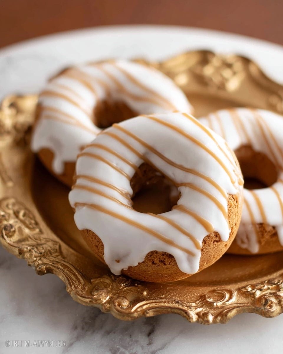 Three donuts sit on an ornate gold plate placed on a white marbled surface. Each donut has a light brown base color with a smooth white glaze that covers the top and slightly drips down the sides. A light brown icing is drizzled in thin stripes over the white glaze, creating a striped pattern. The donuts have a soft and slightly textured look, with a clear hole in the center. Photo taken with an iphone --ar 4:5 --v 7