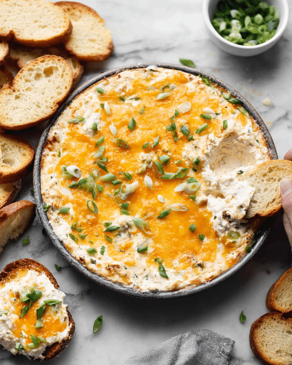A round dish filled with a creamy layered dip, the bottom layer is white and smooth, topped with a melted layer of bright orange cheddar cheese, sprinkled with chopped green onions giving a fresh green color contrast. Around the round dish, there are slices of toasted bread arranged casually on a white marbled surface, some of the dip is spread on one bread slice. A small white bowl filled with more chopped green onions is also visible. The scene includes a woman's hand holding a bread slice partially out of the frame. Photo taken with an iphone --ar 4:5 --v 7