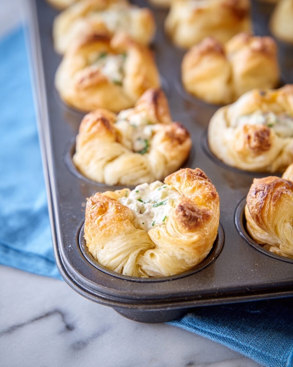 The image shows a close-up of several small pastries in a dark metal muffin tin, each pastry made of flaky, golden-brown puff pastry that is puffed up and slightly open at the top to reveal a creamy white filling with small green herb pieces. The pastries have multiple soft, layered dough folds creating a textured look, and the muffin tin rests on a white marbled surface with a light blue cloth partially visible at the bottom. photo taken with an iphone --ar 4:5 --v 7