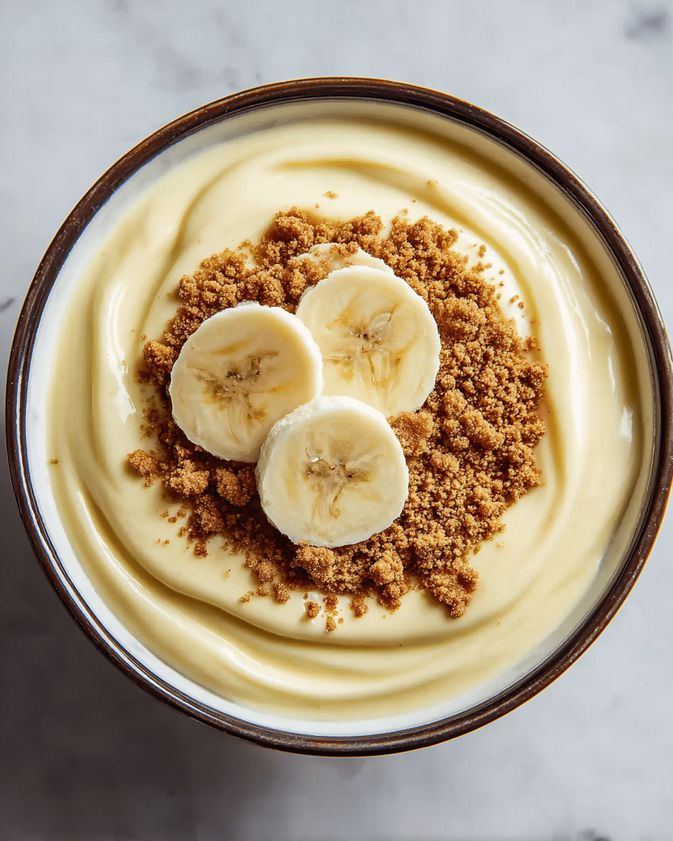 A white bowl with a dark brown rim holds a creamy light yellow pudding with smooth, swirled texture covering the entire bowl. On top, there is a round layer of brown cookie crumbs in the center, forming a circle. Three thick, even slices of pale yellow banana with visible seeds and soft texture are placed on top of the crumbs. The bowl sits on a white marbled surface. photo taken with an iphone --ar 4:5 --v 7