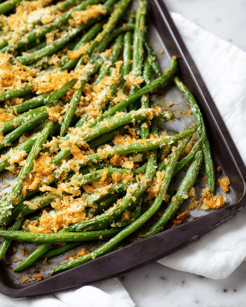 A close-up image of a dark baking tray filled with cooked green beans spread out in a single layer, topped with scattered bits of golden brown and light yellow crispy breadcrumbs and grated cheese, all resting on a white marbled surface with a white cloth underneath part of the tray, showing the green beans slightly wrinkled and roasted. photo taken with an iphone --ar 4:5 --v 7