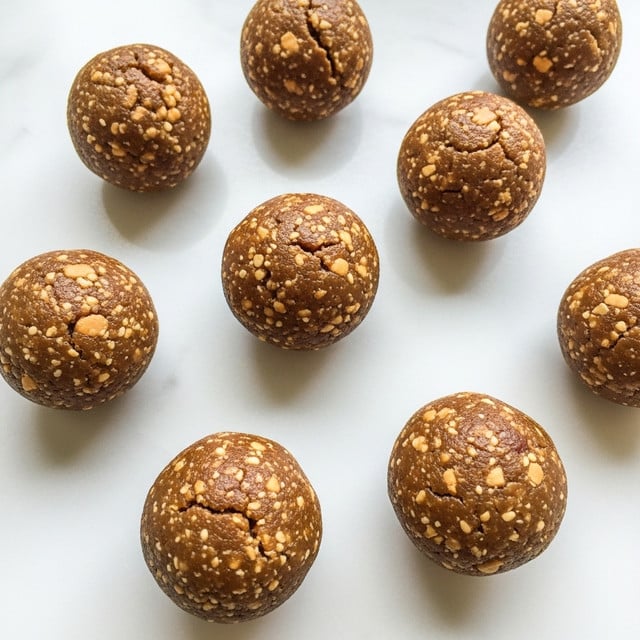 The image shows several round, brown energy balls spread out on a smooth white marbled surface. Each ball has a slightly rough texture with visible small bits inside, indicating a mix of ingredients like nuts or oats. The balls are uniform in size and have a matte finish with some natural cracks and creases on their surfaces. The layout is simple, with the balls placed in an easy and inviting pattern. photo taken with an iphone --ar 4:5 --v 7