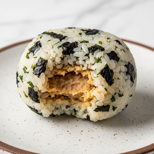 A round rice ball is shown with a bite taken out of it, revealing a soft, light brown filling inside. The outer layer is made of sticky white rice mixed with small pieces of dark green seaweed, which are scattered unevenly throughout the surface. The rice grains appear moist and slightly shiny. The rice ball sits on a white plate with a speckled brown rim, and the background has a white marbled texture. Photo taken with an iphone --ar 4:5 --v 7