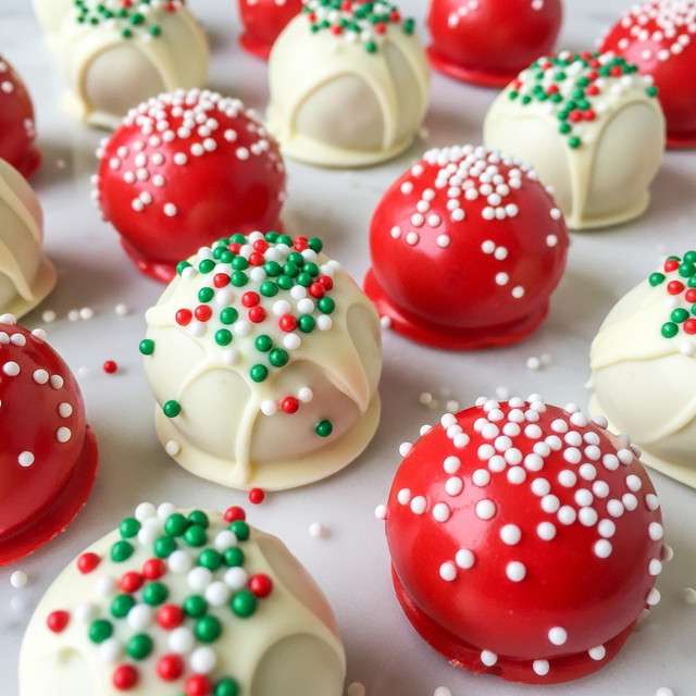 The image shows small round balls covered in smooth, glossy coatings; some are bright white with tiny red, green, and white round sprinkles on top, while others are bright red with white sprinkles. Each ball has a slightly shiny surface and sits on a white marbled texture, scattered randomly. The balls are close together, with a few sprinkles scattered around them, adding a festive look. photo taken with an iphone --ar 4:5 --v 7