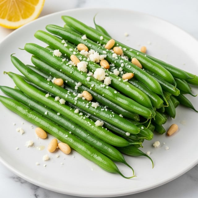 The image shows a white plate filled with bright green cooked green beans stacked in a loose pile. The green beans have a shiny, slightly oily texture and are topped with small bits of white grated cheese and light brown toasted pine nuts scattered unevenly across them. The plate rests on a white marbled surface, and a slice of lemon is partially visible in the top left corner. The overall look is fresh and simple with a mix of smooth green beans, tiny melted cheese flakes, and crunchy pine nuts. Photo taken with an iphone --ar 4:5 --v 7