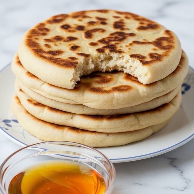 The image shows a stack of four flatbreads with a round shape and light brown color, each bread having a slightly different shade of toasted spots on its surface. The top flatbread has a bite taken out of it, showing a soft and airy texture inside. The flatbreads are layered one on top of the other on a white plate with a subtle blue pattern on the edge. In the foreground, there is a clear glass bowl filled with golden honey, partially visible at the bottom of the image. The scene is set on a white marbled textured surface. photo taken with an iphone --ar 4:5 --v 7