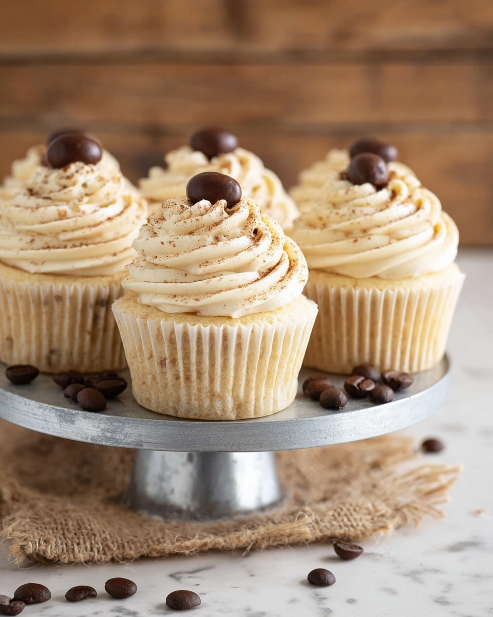 The image shows five vanilla cupcakes placed on a round silver metal stand. Each cupcake has a creamy off-white swirl of frosting piped high in a spiral shape, sprinkled lightly with brown powder. On top of each frosting swirl sits a single dark brown shiny coffee bean. The cupcakes have light yellow cake bases wrapped in pale paper liners. The stand is set on a piece of burlap cloth lying on a white marbled surface, with a few loose dark brown coffee beans scattered around. The background is softly blurred with warm wooden tones visible. photo taken with an iphone --ar 4:5 --v 7