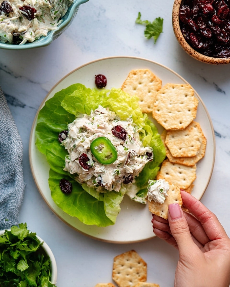 On a white plate placed on a white marbled surface, there is a layer of bright green lettuce leaves forming the base on the left side, topped with a creamy mixture that appears white with small green bits and scattered dark red dried cranberries, finished with a slice of green jalapeño pepper on top. To the right side of the plate, there are several light beige, hexagon-shaped crackers arranged in a small stack and spread slightly outward. A woman's hand is seen holding one cracker dipped in the creamy mixture near the bottom right of the plate. In the background, there is a small white bowl with fresh green cilantro and a light-brown bowl filled with dark red dried cranberries. Photo taken with an iphone --ar 4:5 --v 7