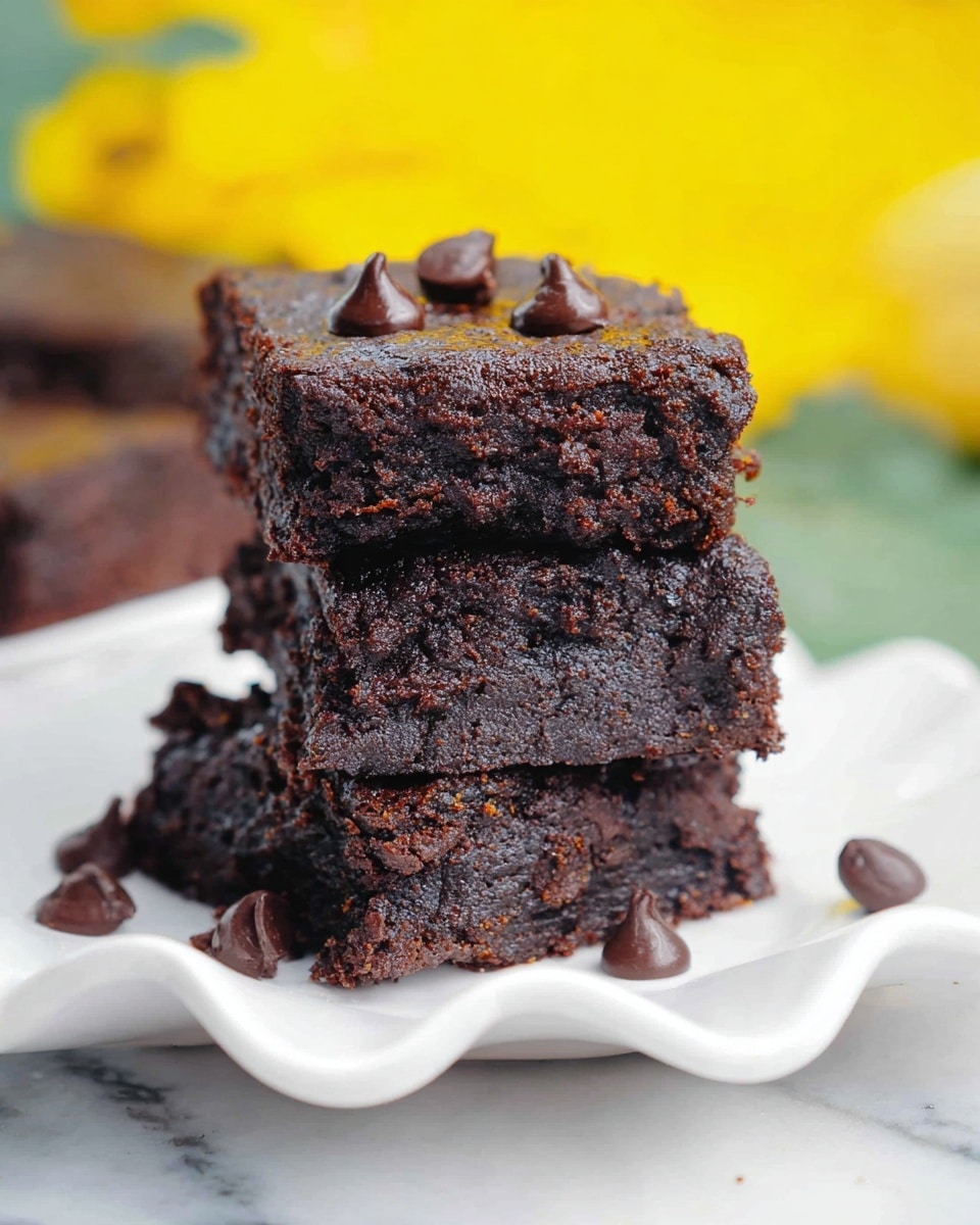A stack of three dark brown brownies with a moist and dense texture are placed on a white plate with a wavy edge. The brownies have small chocolate chips visible inside and a few chocolate chips scattered around the plate. The top brownie has three small, glossy chocolate chips on its surface. The background is blurred with bright yellow and green colors, and the plate sits on a white marbled texture. photo taken with an iphone --ar 4:5 --v 7