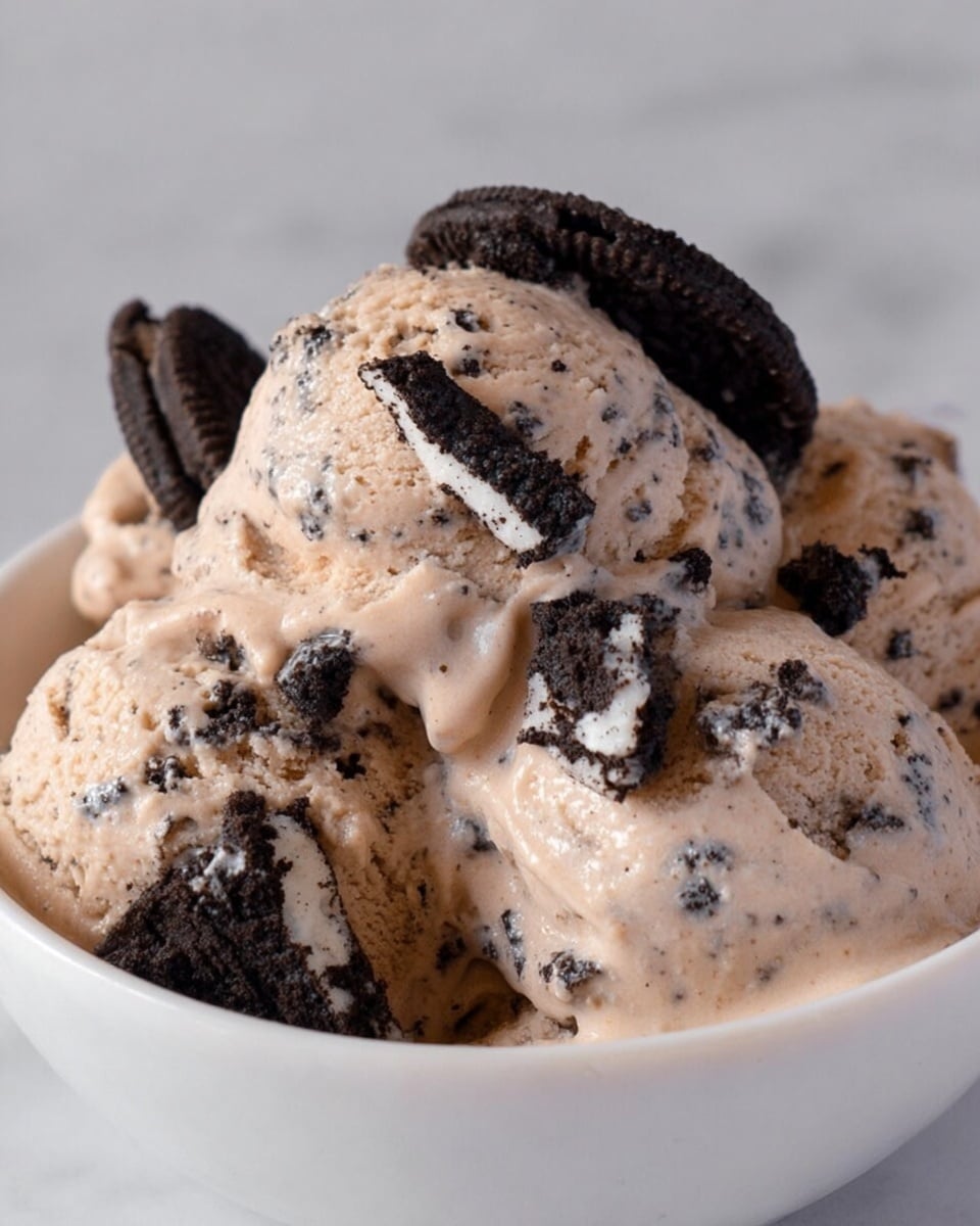 This image shows several scoops of cookies and cream ice cream in a white bowl, placed on a white marbled surface. The ice cream is light brown with dark cookie bits mixed in, creating a speckled texture. On top of the scoops, there are pieces of black sandwich cookies with white cream filling, some broken into smaller chunks and scattered evenly. The ice cream looks creamy and soft with a slightly glossy finish. Photo taken with an iphone --ar 4:5 --v 7