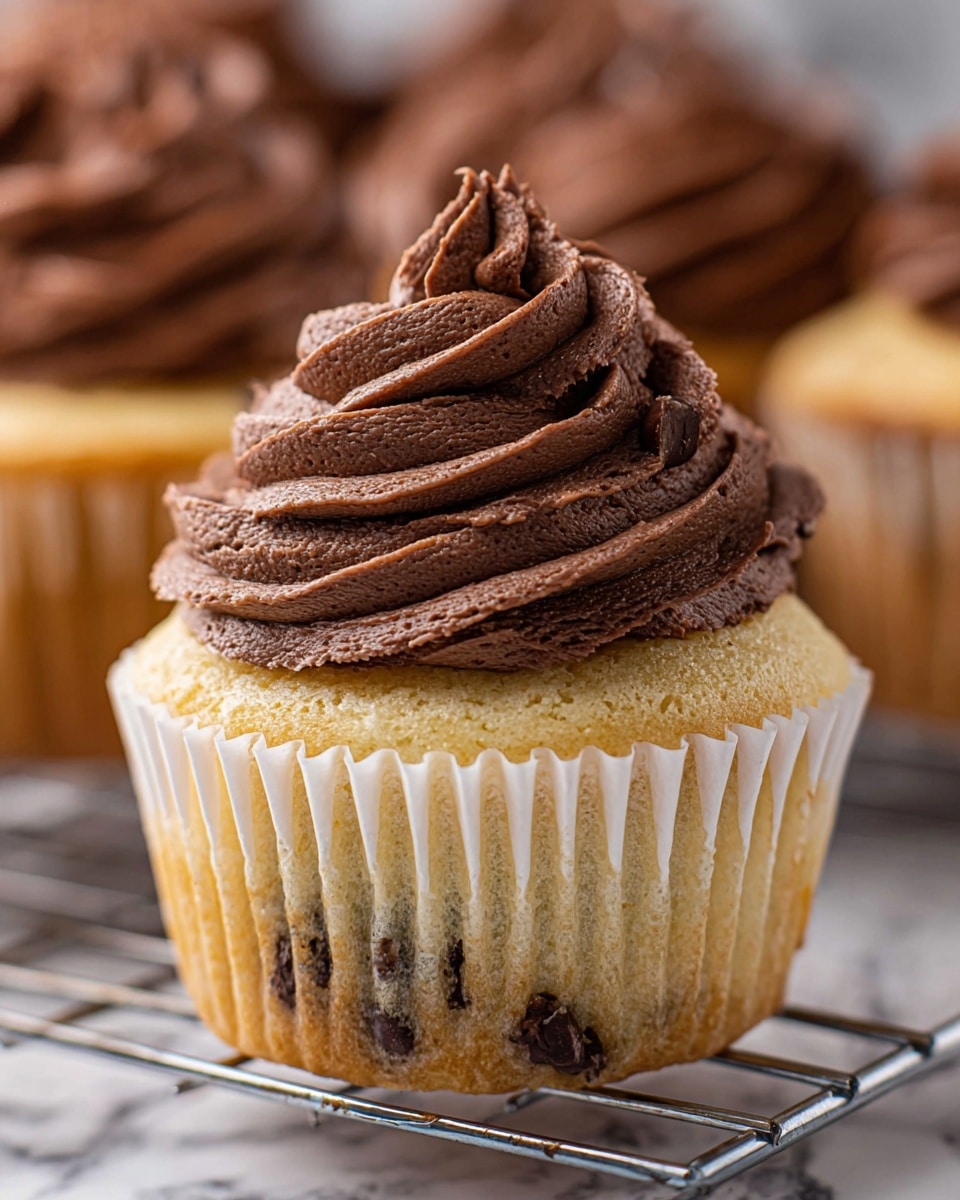 This image shows a close-up of a single cupcake with two layers: the bottom layer is a light golden cupcake with small dark chocolate chips visible inside, wrapped in a white paper liner with gentle folds; the top layer is a thick, creamy, dark chocolate frosting piped in smooth, wide swirls with small air bubbles, forming a tall, textured peak on top of the cupcake. The cupcake sits on a metal cooling rack set on a white marbled surface, with blurred cupcakes in the background. photo taken with an iphone --ar 4:5 --v 7
