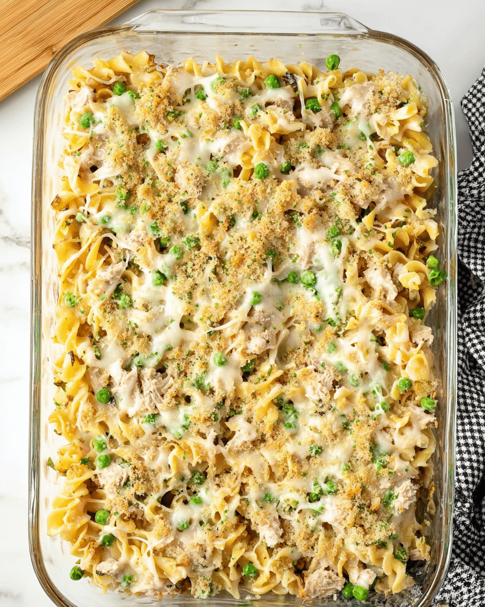 A glass baking dish filled with a layered pasta casserole, showing twisted yellow egg noodles mixed throughout with green peas and chunks of light brown tuna scattered evenly. The top is covered with a thin layer of melted white cheese and light tan breadcrumbs, giving it a slightly crumbly texture. The dish sits on a white marbled surface with part of a light wood board visible at the top left and a black and white checkered cloth at the bottom right. Photo taken with an iphone --ar 4:5 --v 7