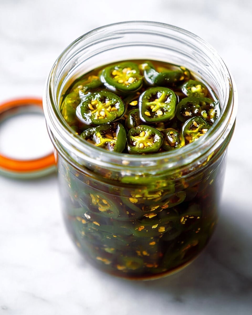 A clear glass jar filled with dark green pickled jalapeño slices soaked in oil, showing the jalapeños' seeds and shiny texture. A metal spoon inside the jar lifts some slices, highlighting their glossy surface and slight wrinkles. The jar rests on a white marbled surface, and some red pepper flakes are visible mixed with the oil. photo taken with an iphone --ar 4:5 --v 7