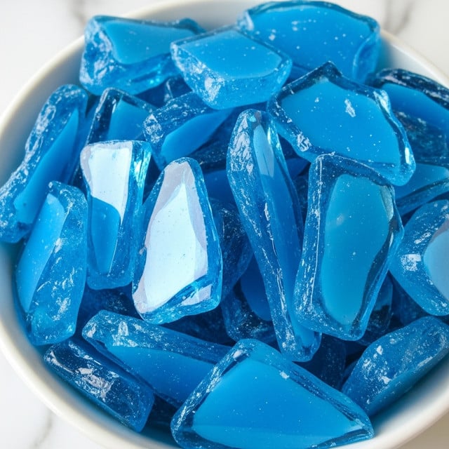 A close-up view of bright blue, shiny shards that look like broken glass piled inside a white bowl. Each shard has sharp edges and smooth, glossy surfaces that reflect light, showing different shades of blue with some areas appearing more transparent or frosted. The shards overlap in a random, uneven way, creating a textured pile that fills the bowl. The bowl sits on a white marbled surface, adding a clean, simple background to highlight the vibrant blue pieces. photo taken with an iphone --ar 4:5 --v 7