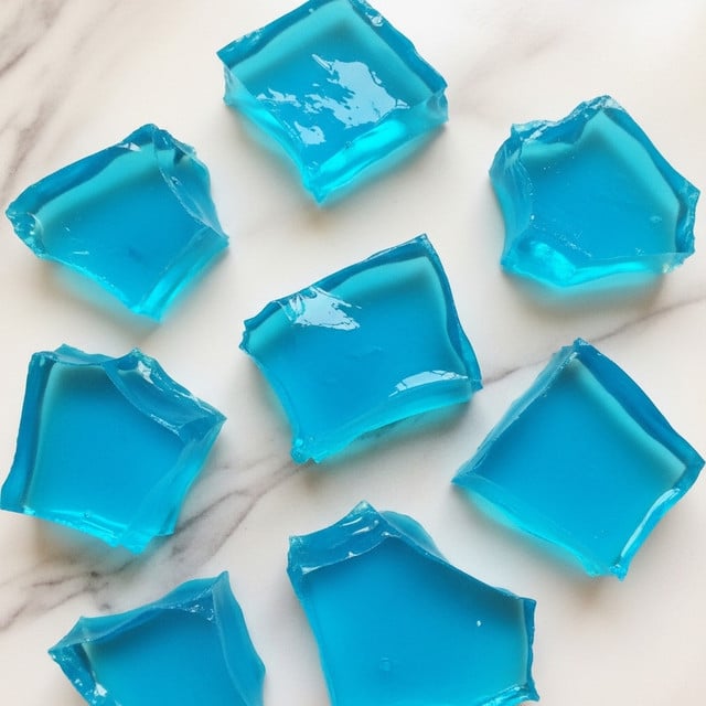 The image shows several broken pieces of bright blue transparent jelly scattered on a white marbled surface. The jelly fragments vary in size and shape, with some having sharp edges and others more irregular forms. The pieces have a glossy, shiny texture that reflects light, making the blue color appear vivid and rich. The background's white marble contrasts with the blue jelly, highlighting its translucence and smooth surface. There are no other objects or layers in the image, just the scattered blue jelly shards. photo taken with an iphone --ar 4:5 --v 7