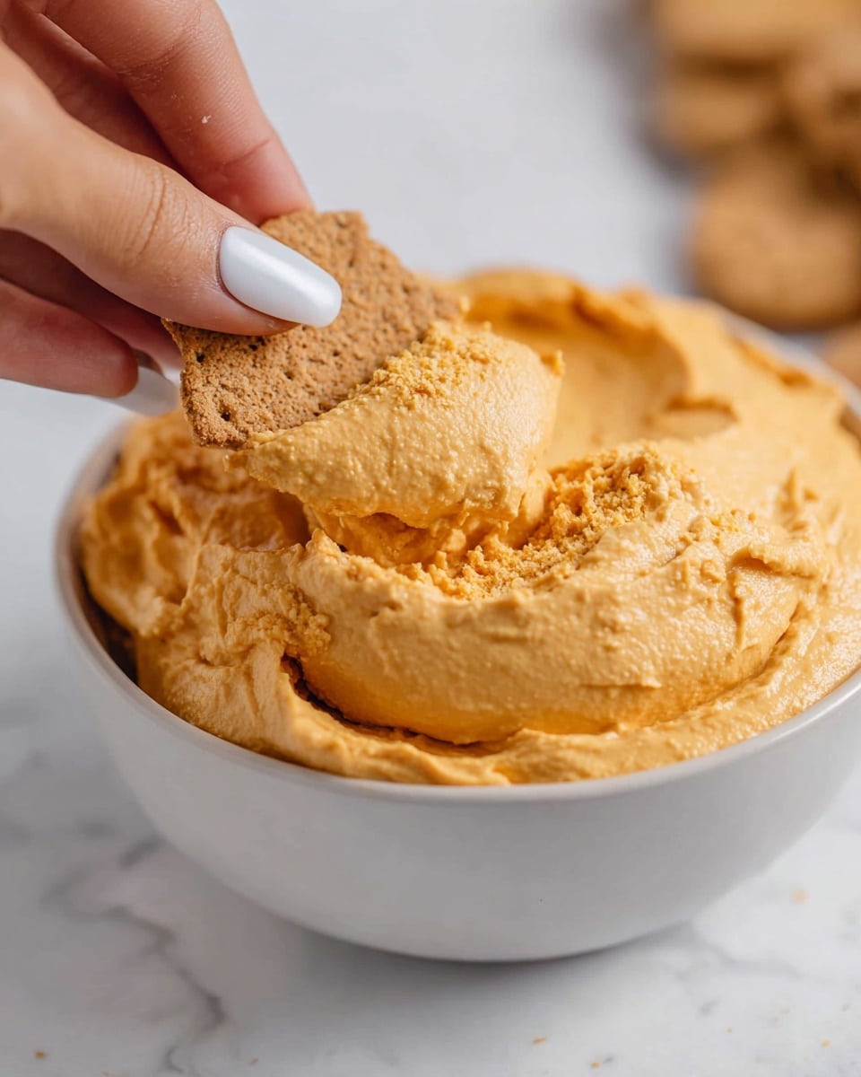 A white bowl filled with a thick, creamy orange dip that has a smooth yet slightly chunky texture. A tan cracker is being dipped into it by a woman's hand with white nail polish, the dip clinging to the cracker in soft, swirled folds. The background is a white marbled surface, giving the scene a clean and bright look. photo taken with an iphone --ar 4:5 --v 7