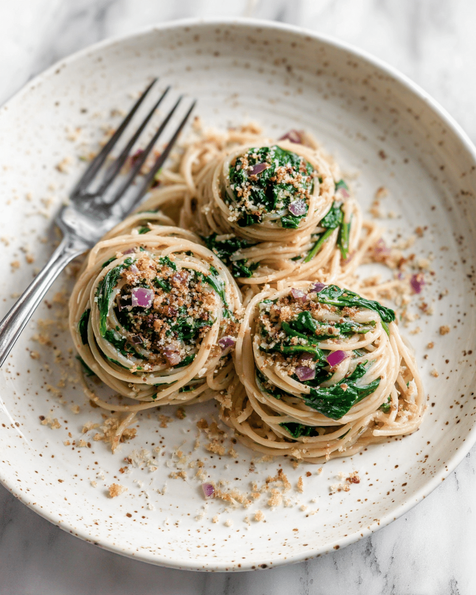 Three small round bundles of spaghetti with green spinach leaves mixed inside sit neatly on a white speckled plate. The creamy beige pasta is sprinkled with tiny bits of light brown breadcrumbs and small pieces of purple onion, giving texture across the dish. A silver fork rests on the plate’s edge, contrasting softly with the plate. The background is a white marbled texture. photo taken with an iphone --ar 4:5 --v 7