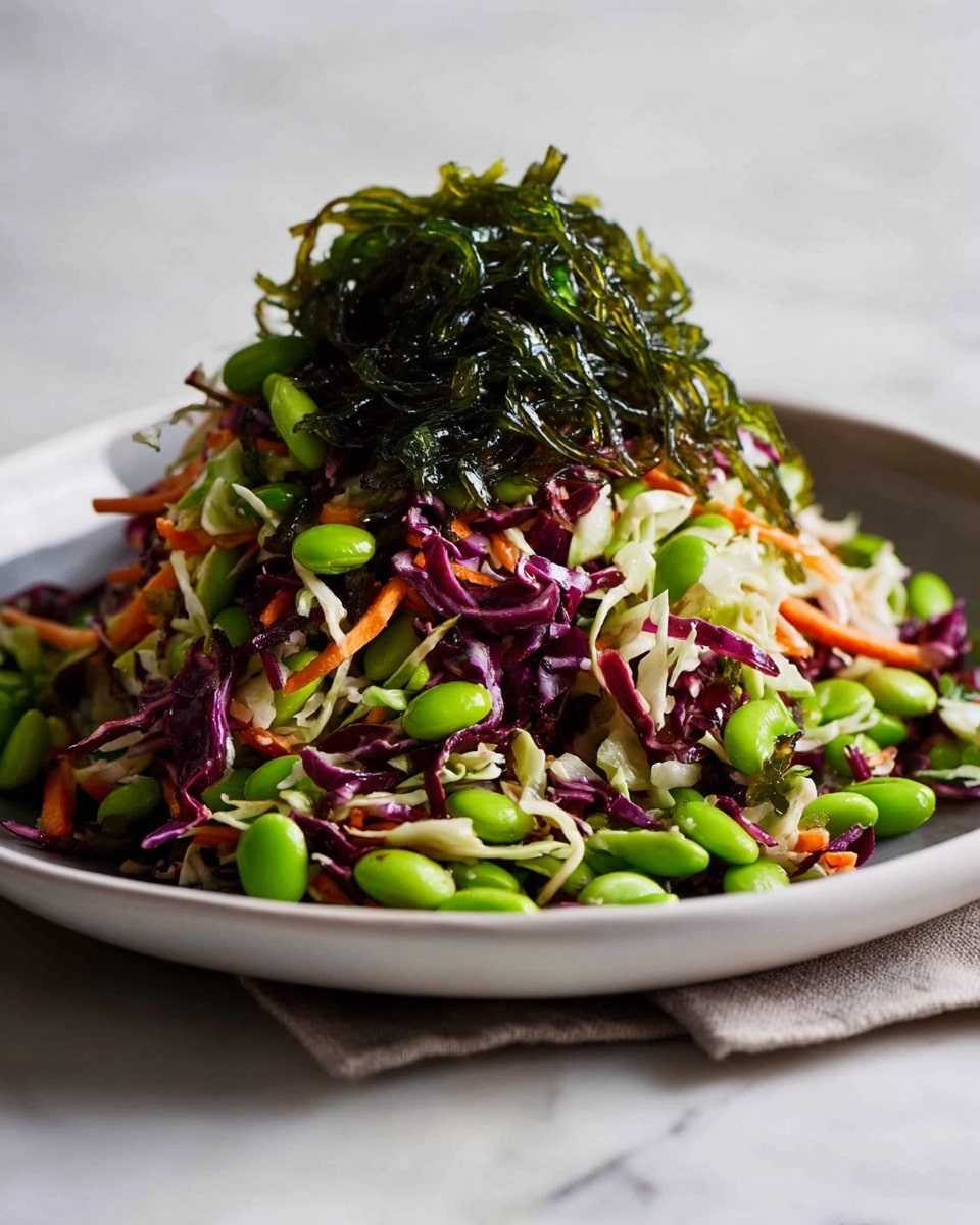 A tall mound of colorful salad sits in a white plate, placed on a white marbled surface. The bottom layer consists of shredded purple and white cabbage mixed with thin orange carrot strips, creating a mix of rough and crisp textures. On top of this base, bright green edamame beans and long, glossy strands of dark green seaweed are spread generously, adding a fresh and glossy look. The combination of vibrant colors and textures makes the salad appear fresh and lively. Photo taken with an iphone --ar 4:5 --v 7