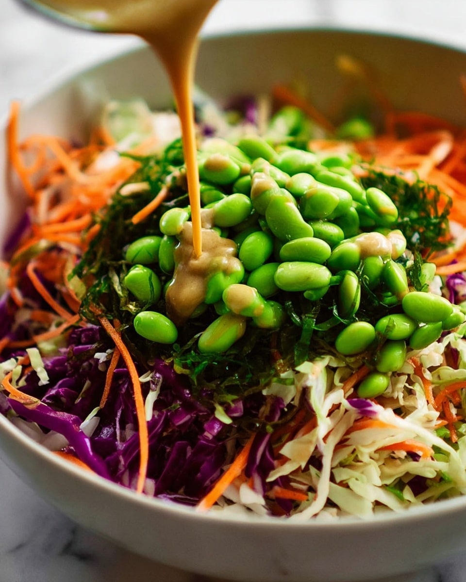 A close-up view of a colorful salad in a white bowl, layered with shredded white cabbage and purple cabbage as the base, mixed with thin orange carrot strips spread evenly throughout. On top, bright green seaweed adds a fresh, shiny texture, while a pile of vibrant green edamame beans sits in the center. A light brown, creamy dressing is being poured in a thin stream over the edamame, adding a smooth texture and contrast to the crisp vegetables. The bowl is placed on a white marbled surface. photo taken with an iphone --ar 4:5 --v 7