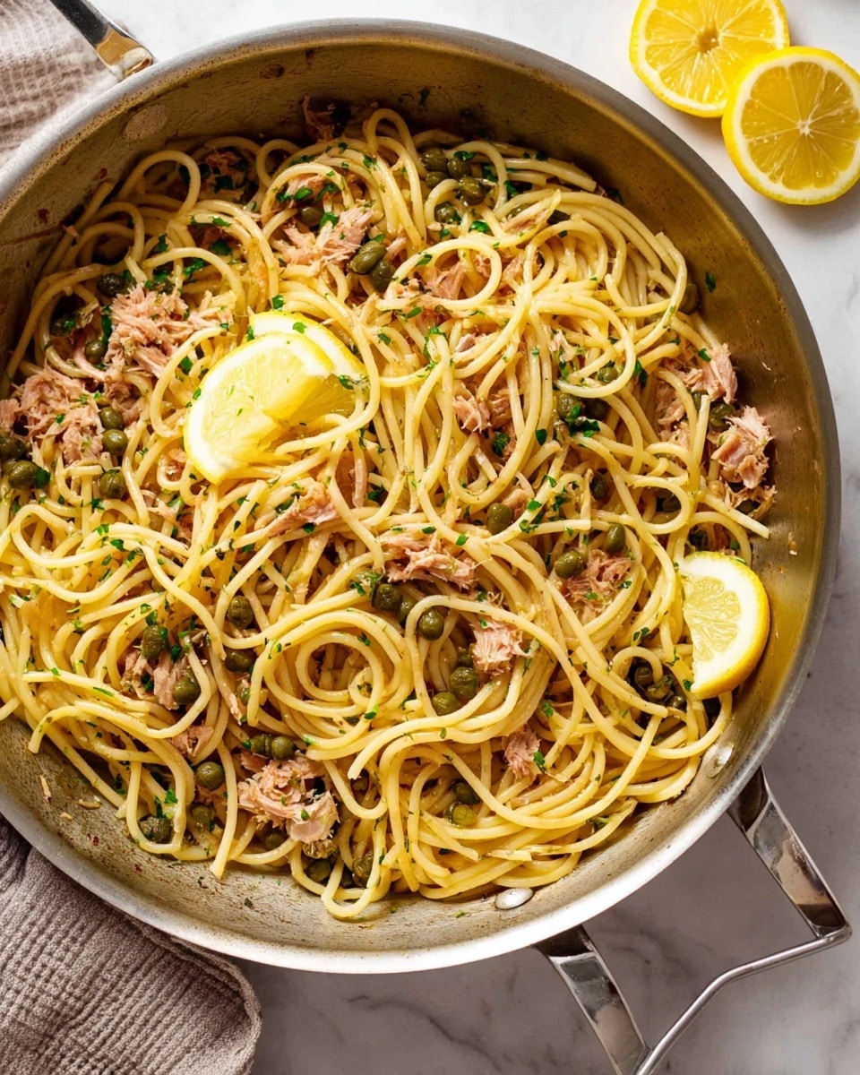 A large silver pan filled with a dish of spaghetti noodles, light yellow in color, mixed with small chunks of light brown tuna, scattered green capers, and finely chopped green herbs. Two thin lemon slices rest on top near the center. The pan sits on a white marbled surface with two lemon slices placed to the right side. The noodles have a glossy, slightly oily texture that shines under the light. photo taken with an iphone --ar 4:5 --v 7