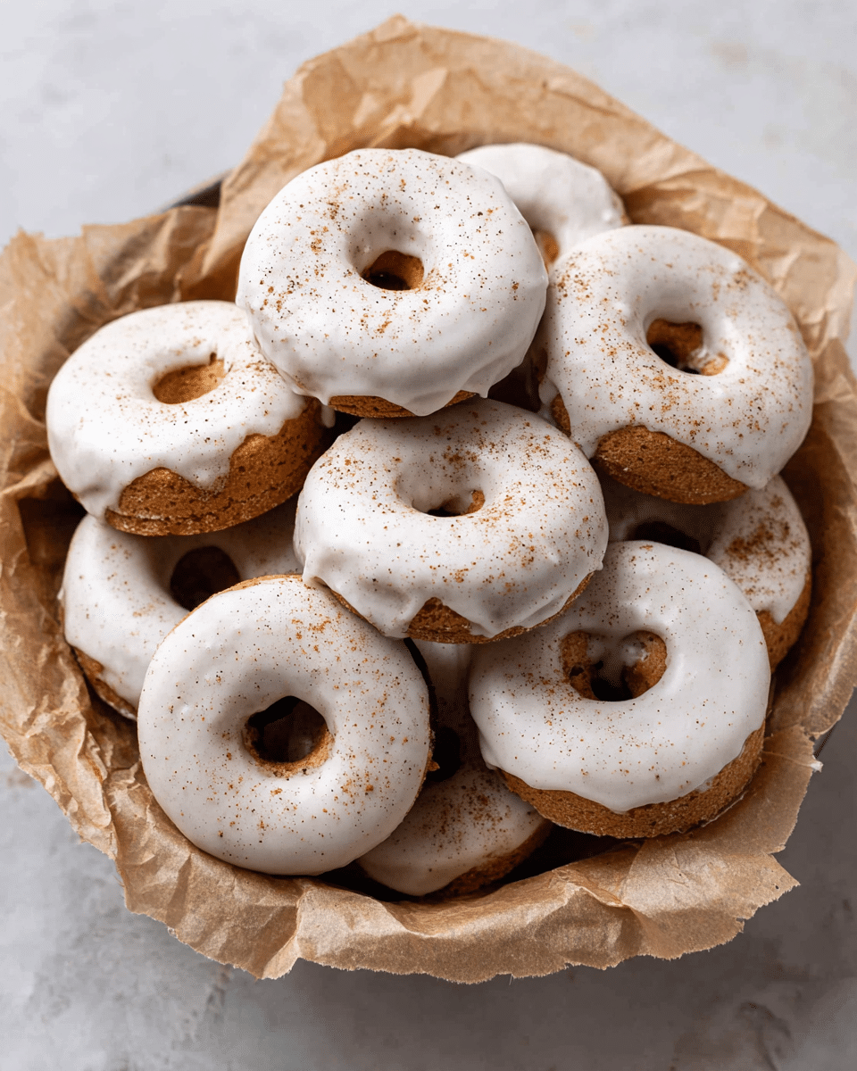 A pile of small round donuts with white icing covers each donut evenly, with a light dusting of brown cinnamon or spice powder on top. The donuts are stacked in a bowl lined with crumpled brown parchment paper. The donuts have a soft, slightly rough brown base visible beneath the smooth white icing layer, and some icing drips slightly down the sides. The bowl sits on a white marbled surface. Photo taken with an iphone --ar 4:5 --v 7