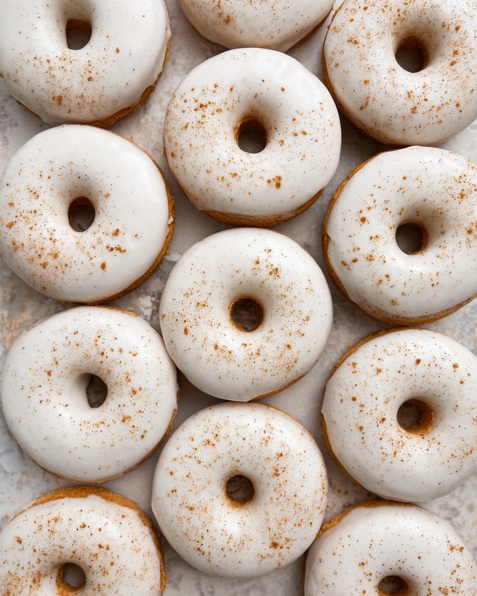 A group of twelve round doughnuts with a central hole are placed closely together on a white marbled textured surface. Each doughnut has one smooth layer of white icing that covers the top and slightly drips over the edges. There is a light sprinkle of fine brown powder scattered evenly across the icing on every doughnut, giving a speckled effect. The doughnuts are golden brown around the edges where the icing does not cover. photo taken with an iphone --ar 4:5 --v 7