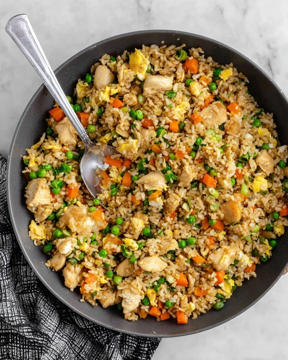 A dark gray pan filled with three visible layers of fried rice ingredients: the first layer is light brown cooked rice with a slightly shiny texture, the second layer consists of bright green peas and small orange diced carrots scattered evenly, and the third layer shows light yellow scrambled eggs and golden brown cooked chicken chunks mixed throughout. A silver spoon sits in the pan's center, partially submerged in the fried rice, all placed on a white marbled surface with a folded black and white checkered cloth near the pan's edge. photo taken with an iphone --ar 4:5 --v 7